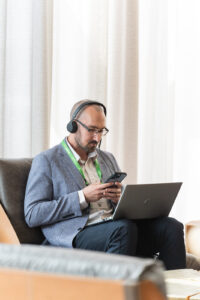 Attendee of Benchmark Gensuite's 2025 subscriber Impact conference, seated at a table with their laptop in front of them. They are wearing headphones and looking at the Benchmark Gensuite mobile app.