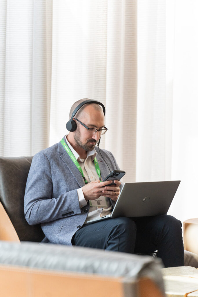 Attendee of Benchmark Gensuite's 2025 subscriber Impact conference, seated at a table with their laptop in front of them. They are wearing headphones and looking at the Benchmark Gensuite mobile app.