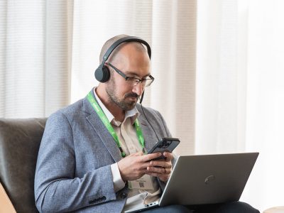 Attendee of Benchmark Gensuite's 2025 subscriber Impact conference, seated at a table with their laptop in front of them. They are wearing headphones and looking at the Benchmark Gensuite mobile app.
