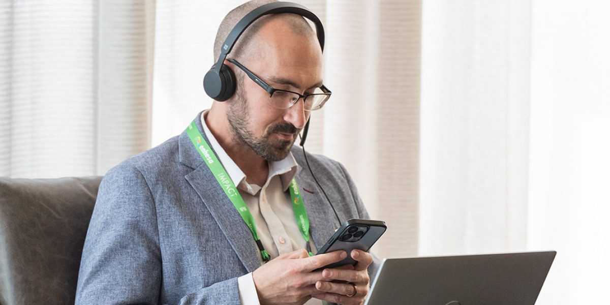Attendee of Benchmark Gensuite's 2025 subscriber Impact conference, seated at a table with their laptop in front of them. They are wearing headphones and looking at the Benchmark Gensuite mobile app.