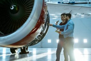 In a Hangar Aircraft Maintenance Engineer Shows Technical Data on Tablet Computer to Airplane Technician, They Diagnose Jet Engine Through Open Hatch. They Stand Near Clean Brand New Plane.